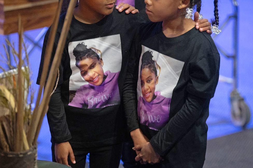 Two girls, whose faces cannot be fully seen, comfort each other at a funeral in Cape Town. They are wearing black with the photo of Esmine Francke in a lilac fleece on the front of their long-sleeved T-shirts. The words 'Being Brave' are written over the fleece - Saturday 30 August 2025.