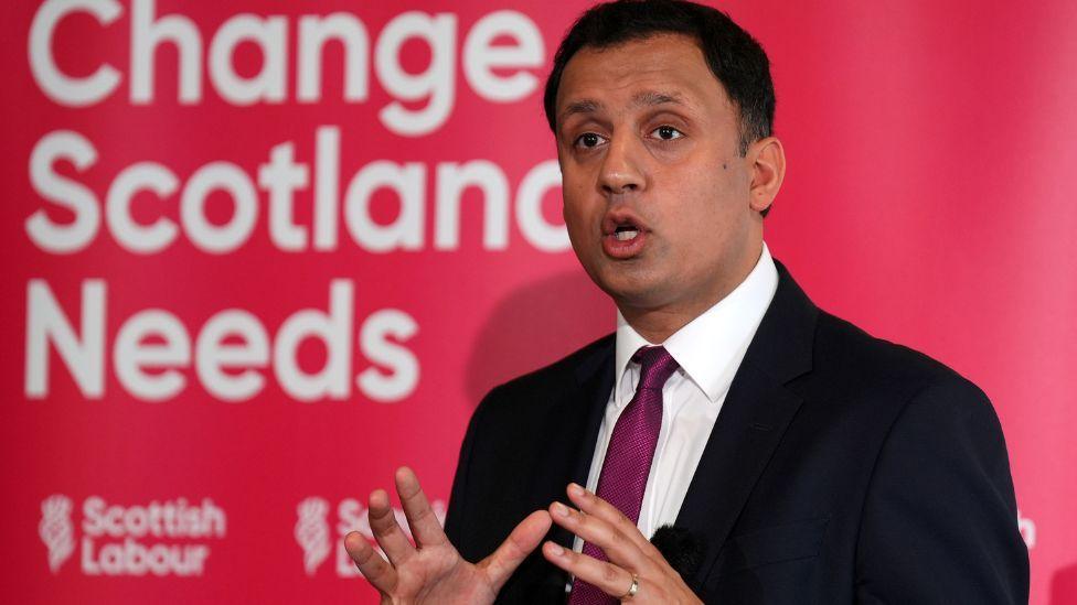 Anas Sarwar, wearing a dark suit and purple tie, speaks in front of a red Scottish Labour sign in a medium close-up shot 