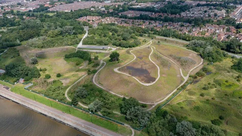 An aerial view of the landfill site where Festival Gardens used to be situated on Liverpool's waterfront. The site is a raised mound surrounded by trees, with homes and streets seen in the distance.