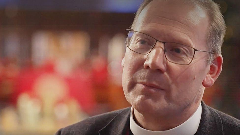 Head and shoulders image of Peter Howell-Jones wearing a dog collar and black jacket. He has short fair hair, greying at the sides, and black wire-rimmed glasses. In the background are blurred candles