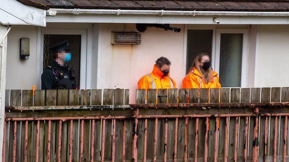 A police officer wearing a face mask stands at a door of a house in Derry, to his left are two people in orange high viz jackets. They are behind a small wooden fence and in front of a white house
