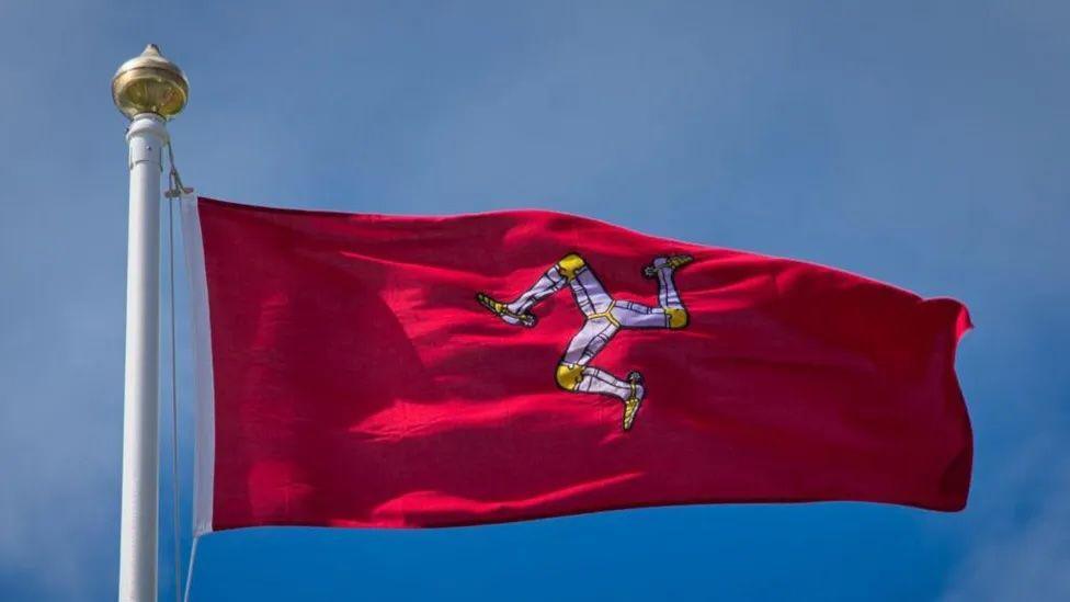 The Manx flag flying at full mast in the wind, it is a red flag, with a white triskellion in the middle and golden knees and feet.