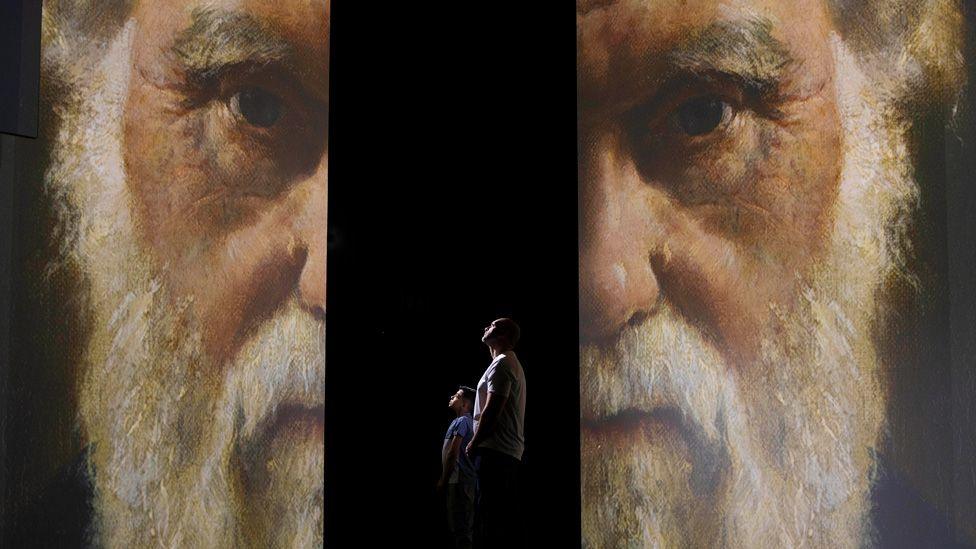 Two halves of Charles Darwin's face on two screens, separated by a space in which a man and boy stand, looking at one of the screens