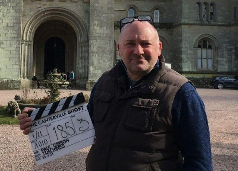 Paul Gibson on a television set, holding a clapper board. He has short grey hair, glasses on the top of his head and is wearing a dark blue long-sleeve top with a brown body warmer over the top. He is stood in front of a large ornate building.