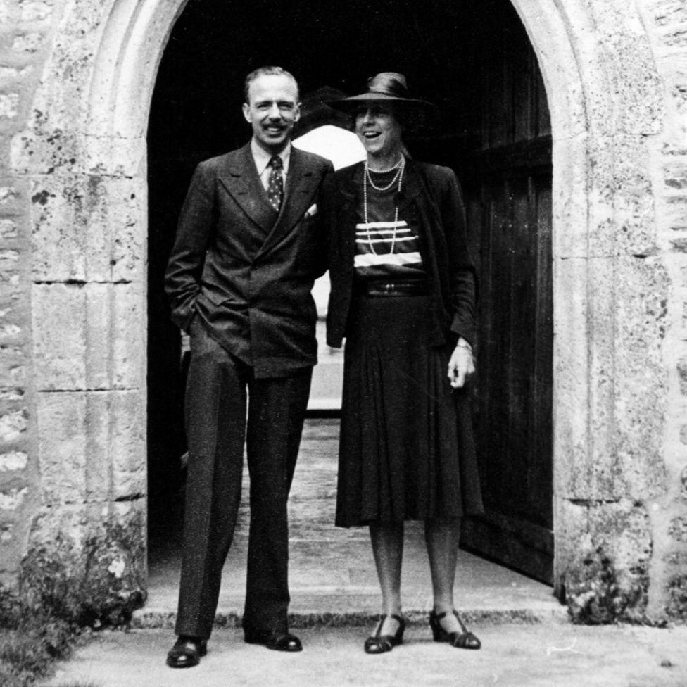 A black and white photo shows a man in a suit and a woman wearing a hat and pearls standing in front of the stone archway at Dartington Hall. They are both smiling.