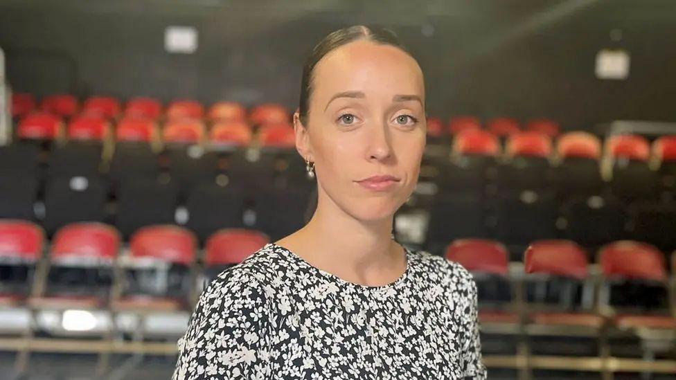 A photo of Ellen Elphick with a pensive, serious expression. She has her hair tied back and is wearing a black and white floral top. There are rows of red theatre chairs in the background 