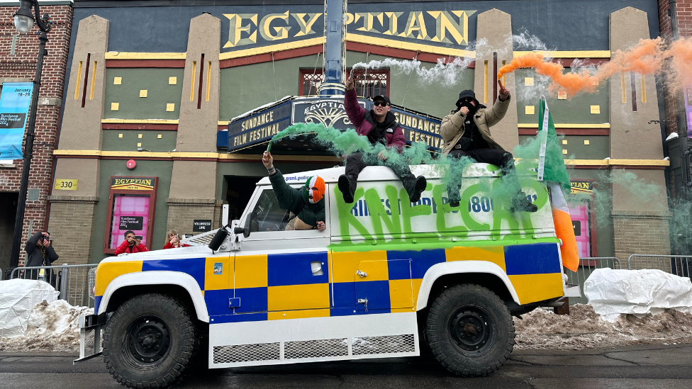 Kneecap members on top of a white land rover, modified to look like a PSNI vehicle, with Kneecap graffiti... two bandmates are sat on the roof letting off green white and orange flares as they pull up to a deco-style Egyptian cinema in Utah, signs advertising Sundance Film Festival are visible and the ground has some snow on it 