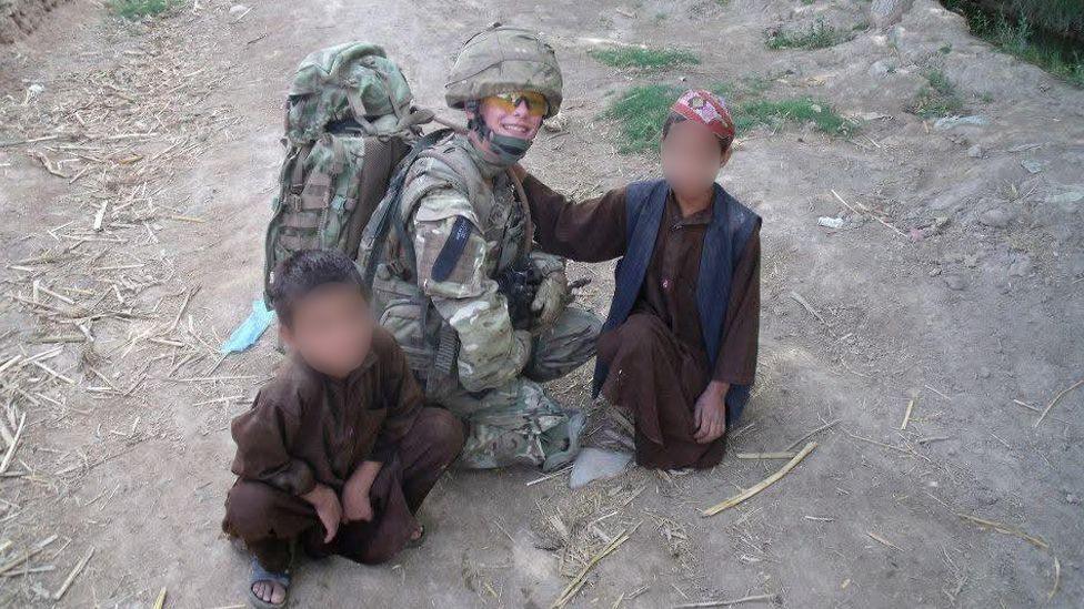 Carol Timone crouching on a dirt track in Afghanistan wearing combat fatigues and a helmet and carrying a large backpack. There are two young boys either side of her wearing traditional Afghan dress. The faces of the two boys have been blurred.