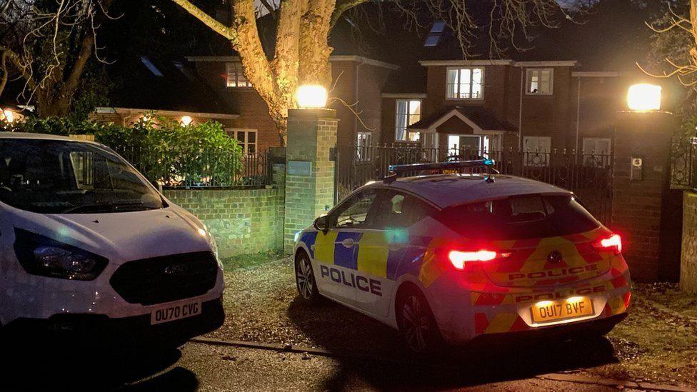 A police car and a police van are parked in a residential area, next to some iron gates. The car's rear lights are on.