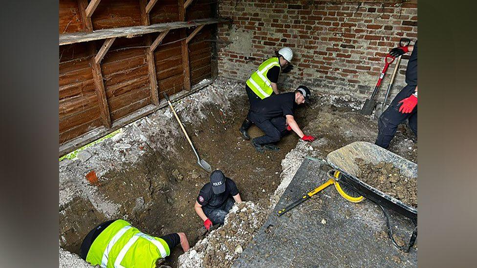 A low angle shot of inside a farm building. Several workers in hi-vis jackets and black tops and trousers dig after a top layer of concrete was removed. 