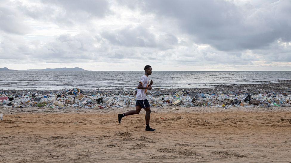 A man running in a white T-shirt, black shorts and black socks jogs along a beach. Behind him can be seen piles of plastic waste washed up by the waves - Conakry, Guinea - Saturday 30 August 2025.