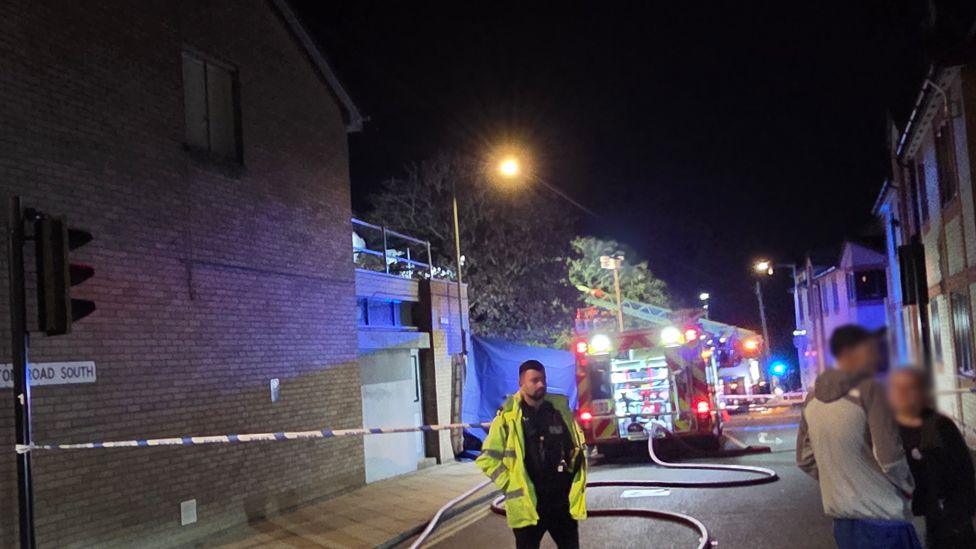 A fire engine is on a street and a long hose is trailing from it down a road. There are brick buildings on either side of the street. A male and a female wearing jackets are standing in the street and there is also a police officer in a yellow hi-vis jacket