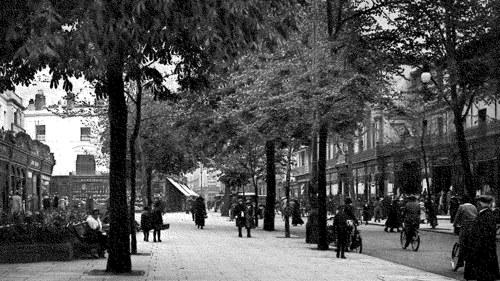 An archived black and white picture of Cheltenham's promenade in 1818. The grainy photo shows the street full of people in Regency clothing with outdoor seating areas and trees lining the street.