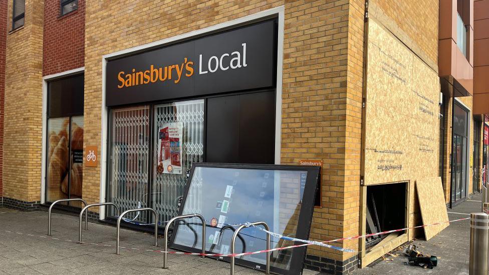 The corner of a Sainsbury's Local convenience store in Milton Keynes. Part of the building has been boarded up after it was damaged. The other side of the building is intact and shows Sainsbury's signage. There is a taped cordon in place around the building and a door which is laying on its side.