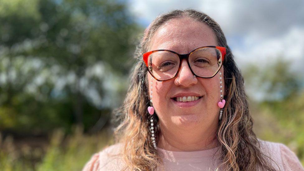 Louise Roe, who is smiling and has long wavy light brown hair and is wearing a large pair of red-tipped glasses, with a chain attached, and a pale pink top. She is standing outside and the background of trees and grasses is out of focus.