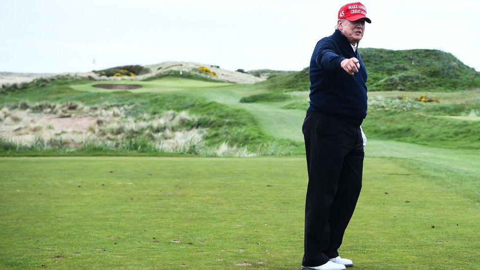 Donald Trump, wearing dark trousers and a dark top, white shoes and a red cap, stands pointing towards the camera on his golf course 