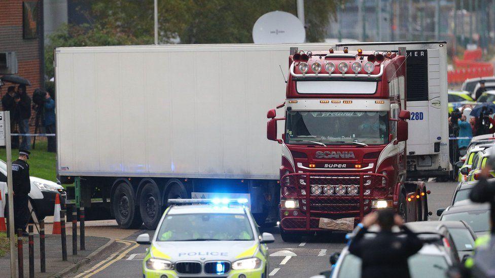 An image of a large red and white lorry discovered in Grays that contained the bodies of 39 Vietnamese nationals. It is turning into a road with a police vehicle in front of it. Cars line one side of the road and several people are taking photographs.