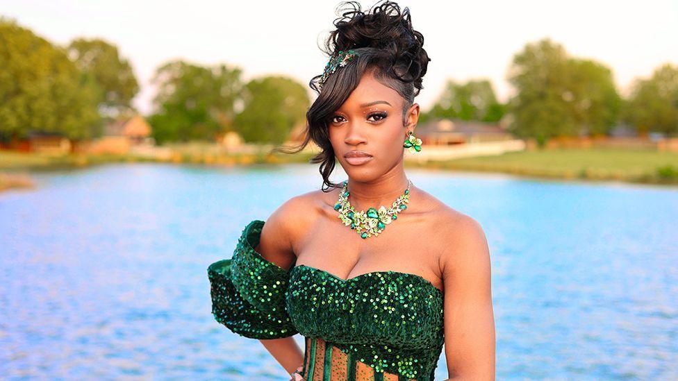 US teenager Trinity Foster wearing a low-cut sequined green dress with a green necklace. She is sitting on the edge of a low wall and behind her is a lake.