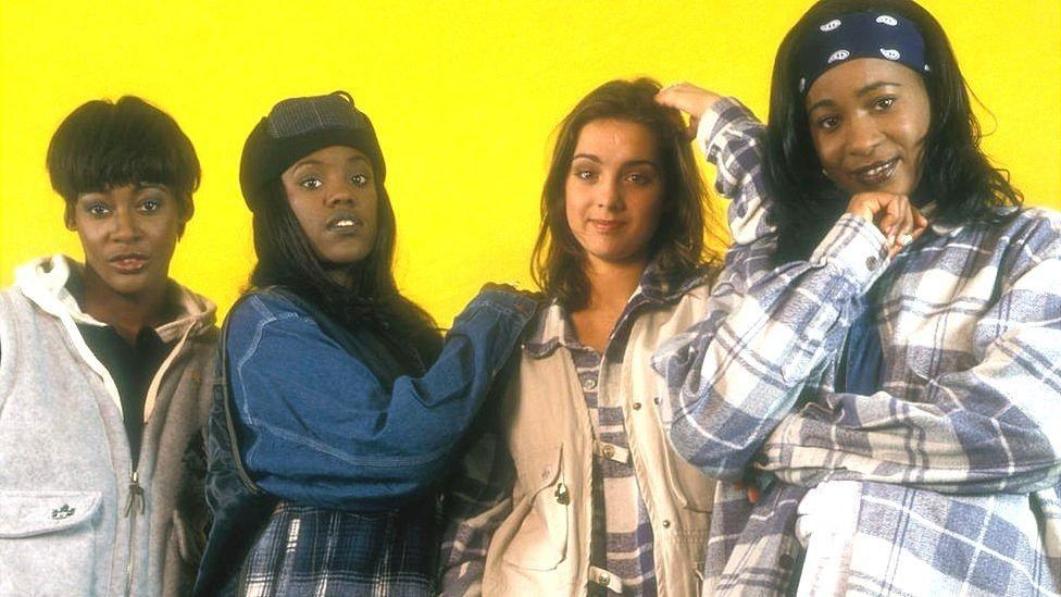 Band members of Eternal Vernie Bennett, Kéllé Bryan, Louise Redknapp and Easther Bennett standing in a line against a yellow backdrop