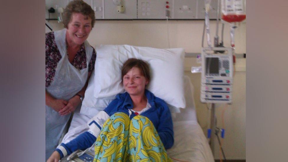A woman is sitting in a hospital bed with a blue long-sleeved top and yellow banana trousers. She has hospital equipment wrapped around her arm. Another woman is standing next to her above the bed in a plastic apron.