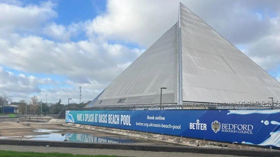 White pyramid-shaped building with grey edging. There is a blue hoarding across the bottom with the slogan MAKE A SPLASH AT OASIS BEACH POOL
There is a car park to the left with a lot of standing water and there are clouds in the sky.