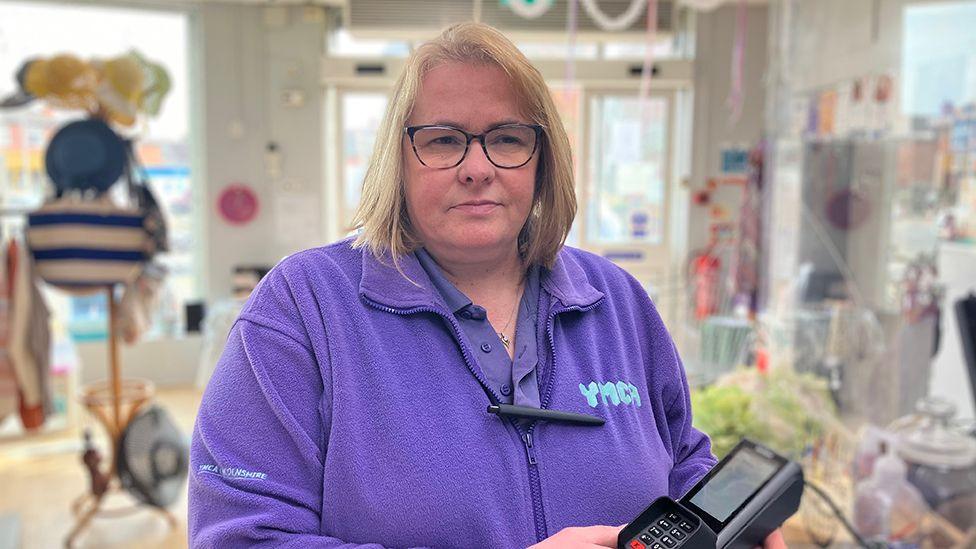 Julie Broughton who has blond hair and glasses and is wearing a purple zip top with the YMCA logo on the front. She is standing in the shop holding a card payment machine 