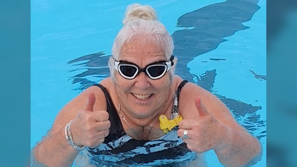 A woman in  a swimming pool wearing dark swimming goggles faces the cameras and raises two thumbs up.