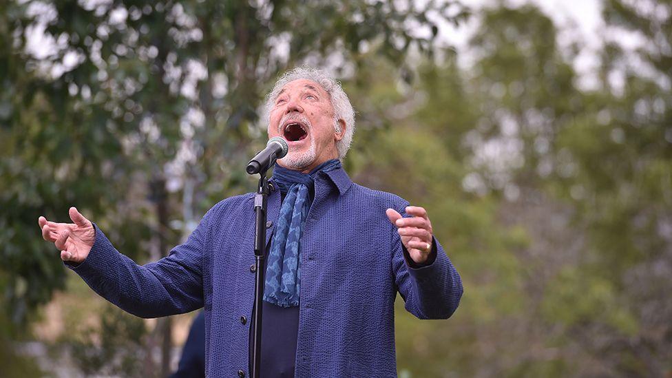 Sir Tom Jones singing with mouth open and arms extended  wearing a blue coat, scarf and shirt 