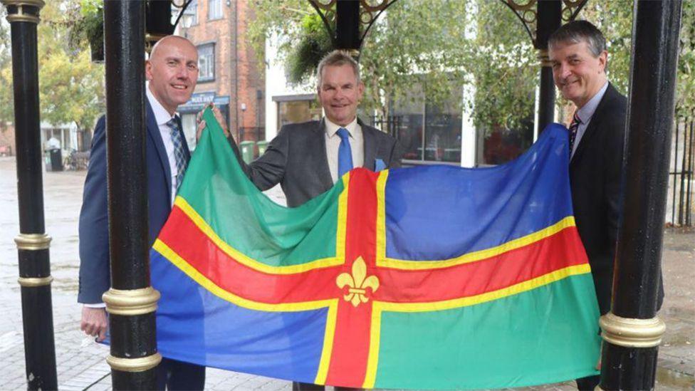 The leaders of Lincolnshire County Council, North Lincolnshire Council and North East Lincolnshire Council holding the county flag, which has a red cross with yellow bands and a yellow fleur-de-lys in the middle, with green and blue quarters. The three men are wearing suits and smiling at the camera.