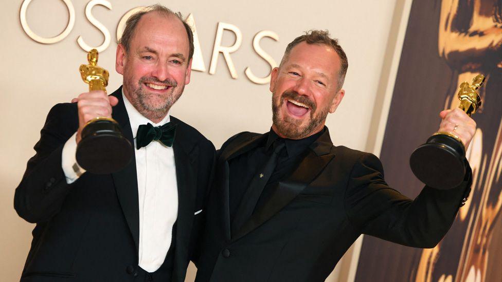 Nathan Crowley (left) and Lee Sandales (right) pose with Oscars. Mr Crowley is wearing a black suit with a white shirt and black bow tie. Mr Sandales is wearing a black suit with a black shirt and a black tie. Both are holding an Oscar in the air and smiling.