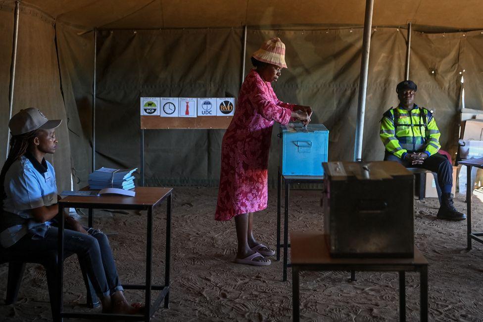 A voter in a pink dress and straw hat casts her ballot as an electoral official sitting to the left and a Botswana Police officer in a yellow jacket uniform sitting on the right, look on. The voting box is on a small table in the centre of a tented polling station. October 30, 2024