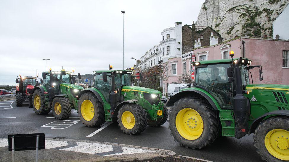 Tractors driving in Dover. They are green and there are cliffs and houses visible in the background. 