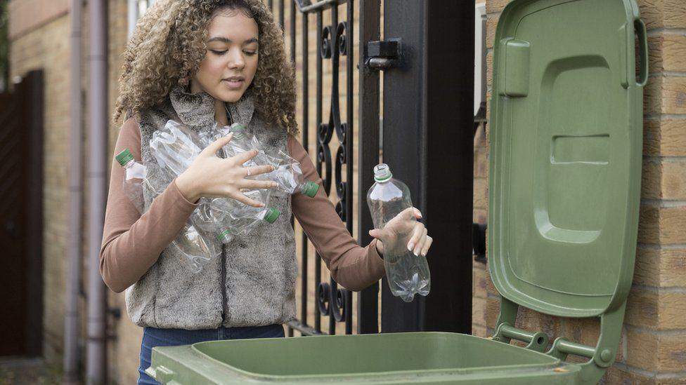 A young woman wearing a brown t-shirt and furry bodywarmer puts a large number of empty plastic bottles into a large green recycling bin.