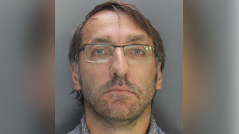 Custody image of Nicholas Stubley who has grey hair and is wearing glasses and a grey and white shirt.