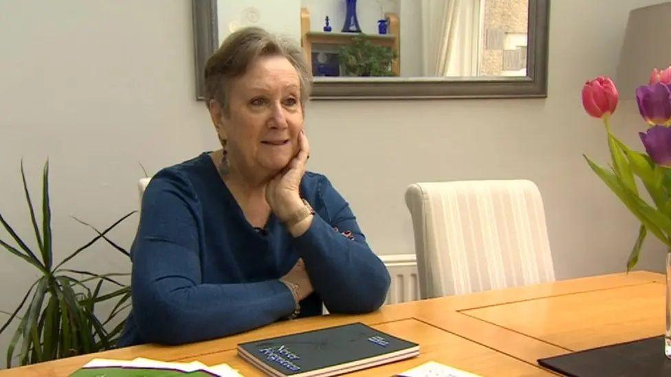 Jane Jackson sits at a dining table with one hand resting on her chin. She has short brown hair and is wearing a dark-blue long-sleeved top.