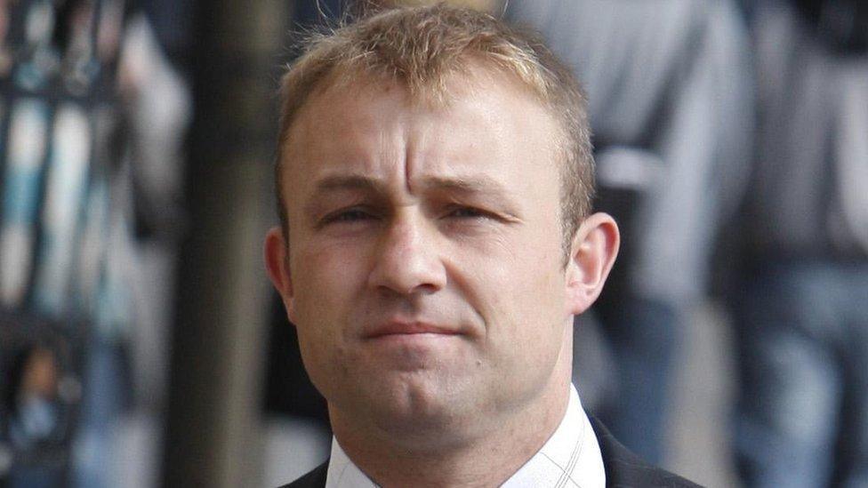 Head shot of a man with short blond hair staring blankly at the camera. He is wearing a dark suit jacket and a white shirt