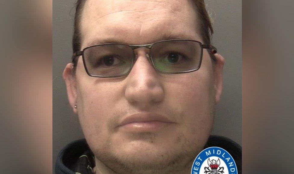 A police mugshot of a man wearing tinted black-frame glasses. He has receding dark brown hair and a stud earring in his right ear.