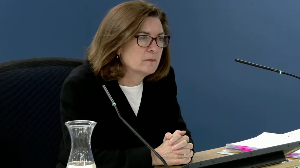 Eluned Morgan sitting behind a desk giving evidence at the inquiry on Wednesday. She is wearing glasses and a black jacket over a white top and has her hands clasped together on the desk as she listens closely to one of the questions she is being asked. She has a pile of notes on the desk to her left hand side and a carafe of water to her right.