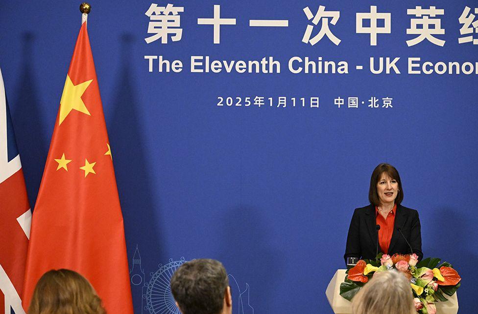 A podium decorated with a floral arrangement stands in front of a blue backdrop displaying Chinese and English text that reads “The Eleventh China-UK Economic…” along with the date and location in Chinese letters. To the left of the podium are two flags: the national flags of China and the United Kingdom. Several people are seated in the foreground, facing the podium.