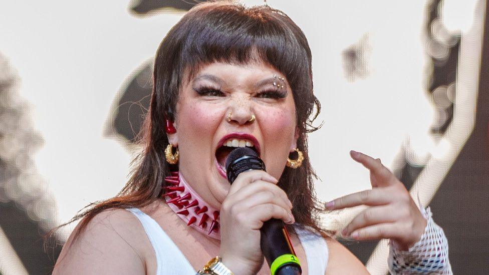 Lola Young singing on stage with her mouth open, one hand holding a microphone and the other gesturing