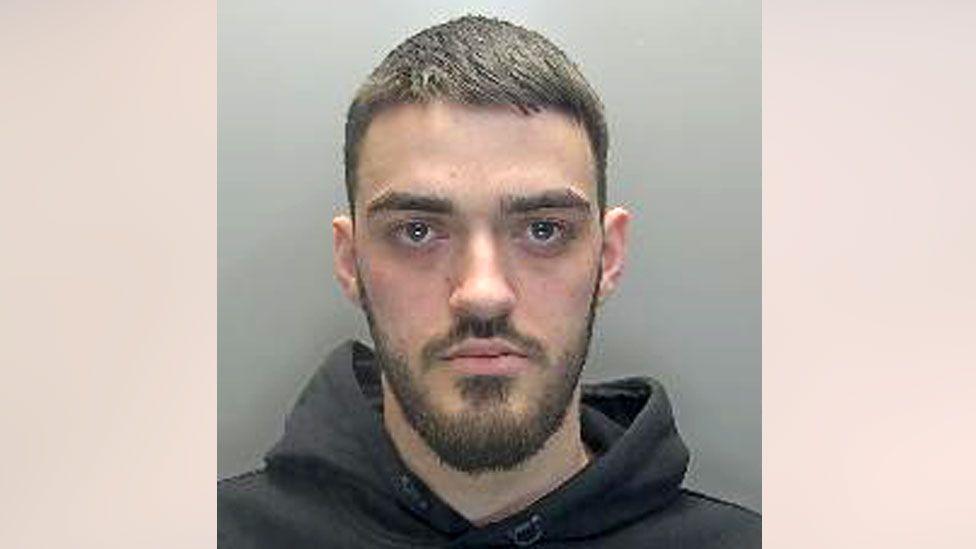 Lirim Shiqerukaj has short brown hair, and a moustache and beard. He is wearing a black hoodie and looking straight at the camera without any expression.