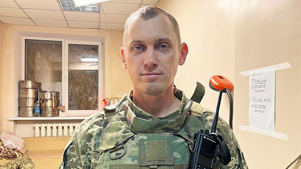 Oleksandr, the head of a medical unit for the Ukrainian army's 25th brigade, photo'd at his place of work, wearing army uniform, and facing the camera in a bare-looking room