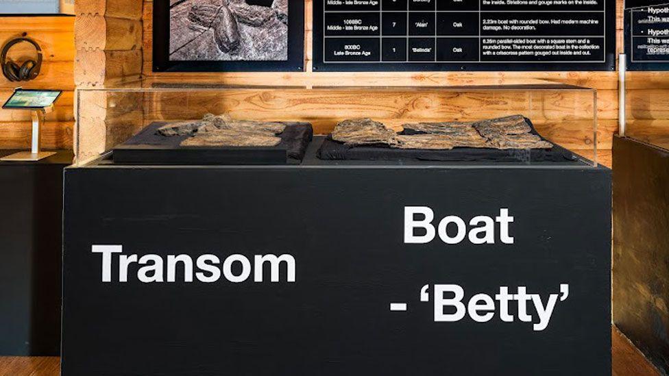 A black display cabinet with transom and boat -  Betty written in white on its front base. Resting on the top, under glass are pieces of ancient wood. The one on the right is a transom board and the larger piece is of a boat. Above it is a black display board with white writing 