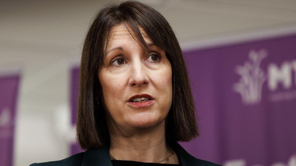 Rachel Reeves, a woman with dark hair parted centrally, in a close up shot. She is visible from the neck up and is looking to her left. She is standing in front of some purple signs, out of focus in the background. 