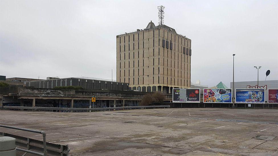 The former Bonny Street police station: a large, square building behind a large, empty, car park with three big billboards to the right