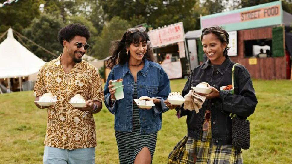 Stock image of three people at a generic food festival, carrying plates of food and drinks