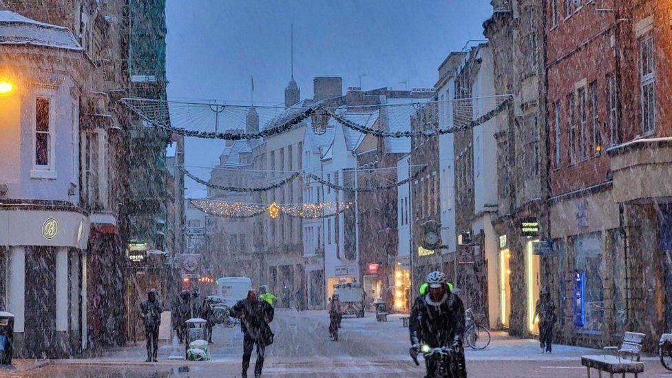 People walking and cycling on road with buildings either side with snow falling
