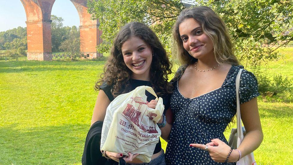 Two women stand in front of a brick viaduct. One of them, with curly dark hair and a black top, holds a bag of shopping with details of a bakery printed on it, while the other, with long brown hair and a dark blue and white patterned dress, smiles and points at the bag