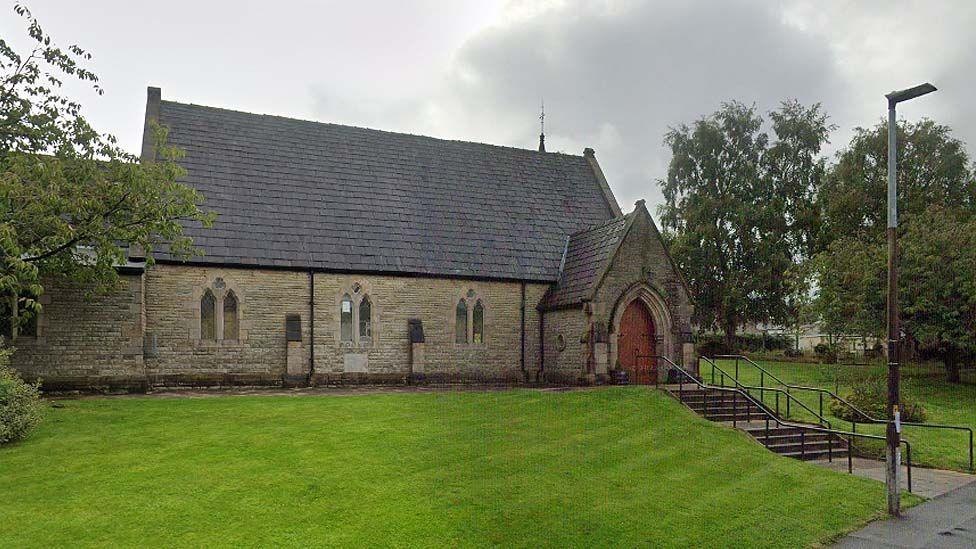 Street view of Trinity United Reformed Church in Brownhill, a traditional looking  one-storey stone church with a large wooden door and grassed areas to the front and side