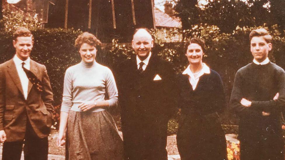 A late 1950s sepia-toned photo of Geoffrey, Sheila, James, Dorothy and Malcolm Webster, with the colour very faded out and all except Malcom smiling. The family are lined up in a garden. Geoffrey and Sheila look late teens, he's wearing a jacket and shirt, with short hair and a tie blowing in the wind; she has short curly hair and is wearing a roll-neck jumper over a tweed skirt, also blowing in the wind. James and Dorothy are middle aged. He's wearing a very dark buttoned up suit over a white shirt and a tie; she is wearing a dark cardigan over  white open necked shirt and dark skirt, with very short hair. Malcolm looks mid-teens and has short hair, his arms are folded over his jumper. 